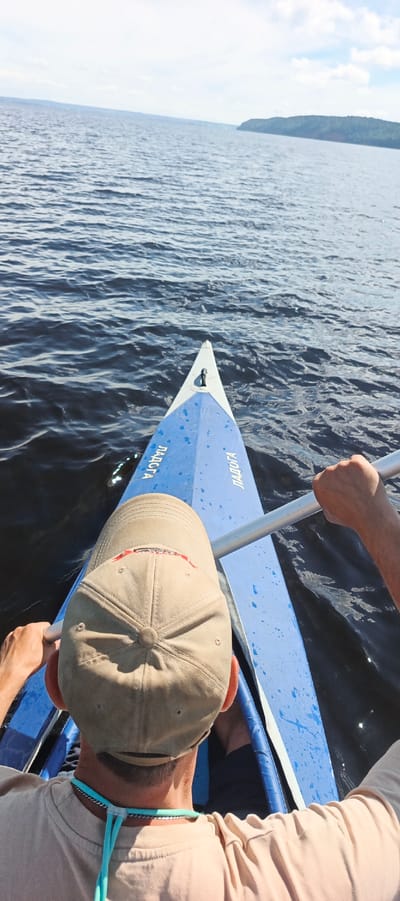 This media file depicts a first-person view from a blue kayak on a large body of water, likely a lake, given the calm surface and distant treed shoreline. The sun is high in the sky, suggesting it is daytime, and the weather appears clear with some scattered clouds visible in the background. The water is a deep blue-gray, with small ripples indicating a gentle breeze.
In the foreground, the back of a person wearing a tan baseball cap is visible. They are wearing a light-colored t-shirt and a turquoise lanyard with a metal chain around their neck. The person is holding a paddle, its silver shaft visible as they appear to be paddling the kayak. Another person's hands are also visible, holding the other end of the paddle.
The kayak itself is blue with white trim and has some water splashes on its surface. Visible text in white, Cyrillic lettering reads "ЛАДОГА" on the sides of the kayak. This text, along with the provided location context of Kostovaty, Russia, suggests the location is Lake Ladoga, the largest lake in Europe, which is situated in northwestern Russia. The overall mood is one of peaceful recreation and enjoyment of the outdoors.