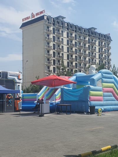 The image shows a bright, sunny day in Fergana City, Uzbekistan, with a clear blue sky and some scattered clouds. The scene is set outdoors, next to a large, multi-story building. To the right of the building, there is a large, inflatable playground structure with bright blue, pink, and green sections. A red umbrella is set up in front of the inflatable, and a few people are visible near a table and chairs, possibly taking a break or supervising. Further to the right, a portion of a street or parking lot is visible, marked with yellow and black bollards and a yellow line. In the foreground, there's a glimpse of a decorated bus or truck with cartoonish figures on its side. The building in the background appears to be a hotel or office building, with many windows and air conditioning units. The text "BESHBOLA BUILDING'S" is visible in red letters on the side of the building. The overall impression is of a lively, possibly recreational, area during the day.