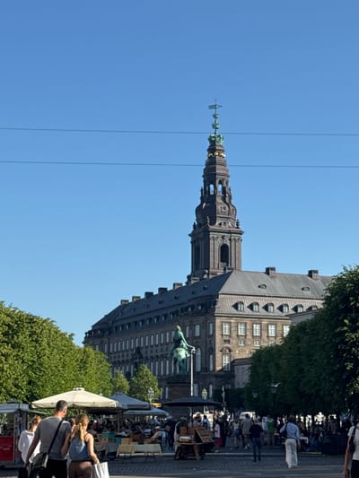 The image depicts a sunny daytime scene in Copenhagen, Denmark, likely in the area of Christiansborg Palace, given the prominent clock tower and building architecture. In the foreground, several people are walking on a cobblestone plaza. A couple is walking away from the camera towards a cluster of outdoor cafes with white umbrellas, where many people are seated and conversing. To the left, a vendor stand is visible.

The midground features a large, imposing building with a grey roof and a dark, ornate clock tower that reaches towards the clear blue sky. A bronze equestrian statue stands in front of the building. Lush green trees line the perimeter of the plaza, partially obscuring the building.

The background consists of the continued expanse of the clear blue sky, with two thin, dark horizontal lines that appear to be wires. The overall atmosphere is one of a lively European city square on a pleasant day.