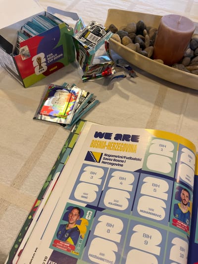 A high-angle shot displays a collection of objects scattered on a surface with a subtle patterned texture. In the top left, a decorative arrangement features a cylindrical brown candle surrounded by light-colored stones within a curved, shallow dish. To the right of this, there's a partially opened sticker album titled "WE ARE BOSNIA-HERZEGOVINA," with spaces designated for player stickers. Several unopened sticker packs are dispersed near the album, alongside loose, empty foil wrappers. A box with "FIFA WORLD CUP 2026" printed on it is in the lower left. The lighting is bright, with a mix of natural and artificial sources. There's no indication of the location being Barrio de Nuevo León, Mexico, nor are there any people visible.