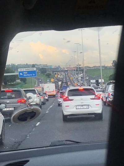 The image displays a multi-lane highway in Sandton, South Africa, exhibiting heavy traffic congestion. The view is from inside a vehicle looking forward through the windshield, which shows minor specks. A circular, dark-colored air freshener or ornament hangs from the rearview mirror, partially obscuring the lower left portion of the view.
A blue overhead road sign in the upper left indicates "124 SANDTON" and "M60 Marlboro Dr" with an arrow pointing left, confirming the location and upcoming exit.
In the immediate foreground, a grey Volkswagen SUV with license plate "CK 57 DV GP" is stationary in the left lane, its brake lights illuminated. To its right, in the middle lane, is a white SUV with a rear-mounted spare tire, also stationary. In the right lane, a white hatchback or small SUV with license plate "LJ 17 LC GP" is stationary, its brake lights illuminated.
The highway extends into the distance, densely packed with various vehicles including cars, SUVs, trucks, and a white bus, all appearing stationary or moving at an extremely slow pace. Rows of tall streetlights with multiple fixtures line the highway median and shoulders.
The sky is overcast with patches of yellowish-orange light visible through the clouds, suggesting either sunrise or sunset conditions. Green foliage is present on embankments and alongside the highway, with distant urban structures visible on the horizon.