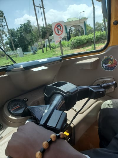 The image provides an interior view from a yellow auto-rickshaw or similar vehicle, looking out through the front window towards a sunny outdoor scene in Vom, Nigeria.

In the foreground, a dark-skinned hand and forearm, adorned with a beaded bracelet featuring light brown/tan and black beads, rests near the vehicle's controls. The bracelet has alternating sets of several light beads and one black bead. Part of the vehicle's black handlebars/steering column is visible, along with a beige dashboard or interior panel. A speedometer gauge is partially visible on the dashboard, displaying "KM/H" and numbers up to 80, with a current reading of "225942" on the odometer. Above the controls, a colorful, tie-dye patterned sticker with the white Nike swoosh and "NIKE" text is affixed to the beige interior trim.

Through the clear window, a bright outdoor scene unfolds under a partly cloudy, blue sky. Prominently in the middle ground is a white building or archway structure with a green, sloping roof and dark arched openings, possibly an entrance gate. Lush green trees and foliage surround this structure and extend across the landscape. In the foreground of the outdoor view, a white "NO PARKING" sign (a red circle with a diagonal line over a black 'P', with "NO PARKI" visible) is partially obscured by the window frame. Several wooden utility poles with wires crisscross the view, suggesting a developed area.

The time of day appears to be bright daylight, indicated by the clear sky and strong shadows. The scene suggests the vehicle is either stationary or moving slowly, providing a clear view of the surrounding environment.
