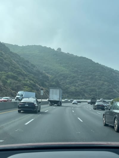 This is a highway scene in Los Angeles, California, United States of America. The image captures traffic on a multi-lane freeway with a steep, tree-covered hillside on the right side. The sky is overcast, creating a hazy atmosphere.
Several vehicles are visible on the highway, including cars, trucks, and a recreational vehicle. In the foreground, a dark-colored sedan is in the right lane, and a red sports car is in the far left lane. A white semi-truck is in the center lanes, followed by other vehicles. A black Lexus sedan is in the lane closest to the camera. A sign indicating "CARPOOL LANE" is visible on the right side of the freeway.
The hillside is densely covered with green vegetation, suggesting a natural environment adjacent to the urban infrastructure. The road surface appears to be asphalt, with clear lane markings. The overall impression is of a busy commute on a major artery in Los Angeles, with the natural landscape providing a backdrop.