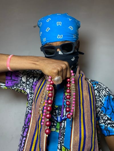 A single individual is depicted from the upper torso upwards, standing against a plain, light-colored wall in Ngleshi Amanfro, Ghana. The person's face is obscured by a black bandana covering the nose and mouth, and black square-framed sunglasses covering the eyes. A blue bandana with white patterns covers the head. The individual wears a multi-patterned shirt, possibly an African print, featuring vibrant blues, purples, greens, and blacks. Layered over this is a woven garment, possibly kente cloth, displaying vertical stripes of brown, blue, and yellow. A pink silicone wristband with text, including "OPM" and "HERE," is visible on the right wrist. The individual's right hand holds multiple strands of beads and necklaces, comprising two strands of large, matte pink spherical beads and two strands of clear, faceted beads. A large, spherical, textured metallic-colored ring is worn on the right index finger. The person's left arm is bent, with the elbow visible on the right side of the frame.