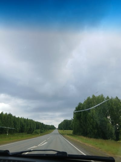 The image shows a view from inside a vehicle traveling on a paved road. The road is straight and asphalt, with white lane markings visible. On either side of the road are grassy verges and dense green trees. The sky above is overcast with a gradient of blue and grey clouds. In the foreground, the dashboard of the vehicle and a portion of the windshield wipers are visible. The scene appears to be in a rural or suburban area, possibly along a highway or a main road outside of a populated center. There are no people or other vehicles immediately visible in the frame. The overall impression is one of a cloudy day journey through a tree-lined landscape.