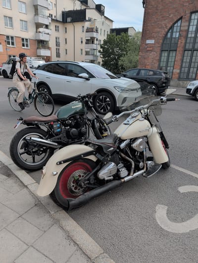 The image depicts a street scene in Stockholm, Sweden, likely in the Norrmalm district, near the intersection of Kungsgatan and Sveavägen, based on the architectural style and street markings. In the foreground, a cream-colored Harley-Davidson motorcycle is parked, its chrome accents glinting. Behind it, a dark green Kawasaki motorcycle is also parked. To the left of the motorcycles, a person wearing a helmet and a backpack is riding a bicycle on the sidewalk. The cyclist is dressed in neutral-colored clothing. In the background, several cars are parked and moving on the street, including a white electric SUV. The buildings in the background are typical of Stockholm's urban architecture, with apartment buildings and a brick structure with large arched windows. The street is paved, and a sidewalk is visible. The overall impression is a bustling urban environment.