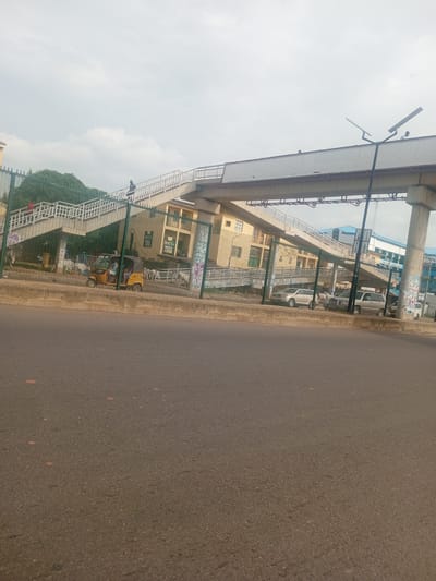 This media file depicts an elevated pedestrian walkway passing over a street in Unguwar Sarki, Nigeria. The walkway is constructed with concrete and metal railings, with a modern, wide overhead bridge structure extending from it. The walkway itself appears to have a gentle slope, indicating it is designed to be accessible. Several individuals are visible on the walkway, some walking, suggesting normal daily activity.

Beneath the walkway, a yellow auto-rickshaw, a common mode of transport in the region, is parked. Behind the auto-rickshaw, several cars are parked along the roadside, including SUVs and sedans, suggesting a commercial or residential area. A chain-link fence separates the street from the area under the walkway. On the building behind the fence, there are signs with text, though they are difficult to read clearly. One sign appears to have "helgia" in prominent lettering, and another smaller text may indicate a business or service. The architecture of the building is utilitarian, with visible windows and what appear to be multiple floors.

The sky is overcast, suggesting a cloudy or hazy day. The lighting is diffused, indicating it is likely daytime, but not a bright, sunny period. The overall scene conveys a sense of urban infrastructure and daily life in Nigeria, with a focus on pedestrian access and common transportation methods. The presence of the elevated walkway and the parked vehicles paints a picture of a busy, functional urban environment.