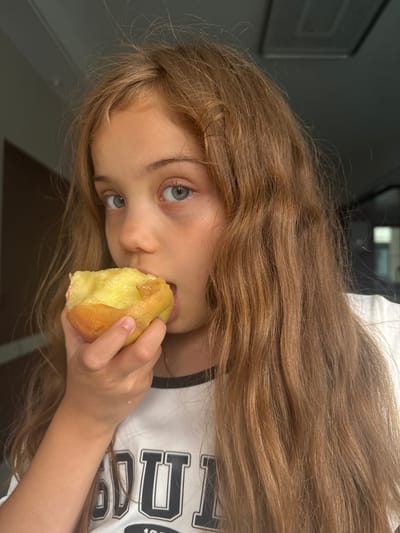 A young girl with long, light brown hair is shown in a close-up, head-and-shoulders shot, looking towards the camera. She is in the process of taking a bite out of a piece of light yellow fruit, which appears to be an apple or pear. Her eyes are light blue and her expression is curious or perhaps slightly mischievous. She is wearing a white t-shirt with black trim and what appears to be lettering in black. The background is out of focus, but suggests an indoor setting with some muted colors and a hint of a framed picture on the wall. The lighting appears to be natural, possibly from a window, and the time of day is indeterminate. There is no visible text in the image that can be read.