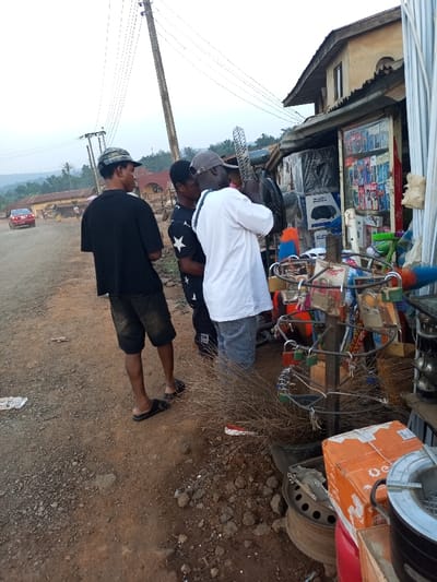 The image depicts a vibrant roadside scene in a semi-urban or rural setting, characteristic of Aramoko-Ekiti, Nigeria, likely in the late afternoon or early evening given the soft, diffused light. Three men are the primary subjects, standing near an open-fronted shop or stall.

The man on the far left wears a black t-shirt, dark shorts, and a camouflage cap, looking away from the camera towards a dirt road where a red car is visible in the distance. The other two men are positioned closer to the shop. The central figure wears a black t-shirt with white star patterns, while the man on the right, wearing a white t-shirt and a grey cap, appears to be examining merchandise displayed at the stall. He holds an item up, possibly a fan or a metallic object.

The shop has a rustic appearance with a corrugated metal roof and shelves. Its storefront showcases a variety of electronics, including mobile phones, visible through a glass display case. Outside, numerous goods are exhibited: a metal stand holds various padlocks, some clearly branded "X-FORCE." Other items for sale include brooms, white pipes, fans, and what appear to be general household wares. A cardboard box, possibly with "GE" branding, and discarded tire rims are visible on the ground near the shop.

The ground is a rough dirt path with scattered pebbles. Overhead, multiple power lines crisscross the sky, supported by a prominent utility pole. The background features distant hills or mountains under a pale, hazy sky, adding depth to the local street scene.