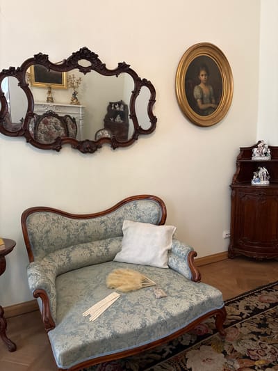 The image depicts a richly decorated interior, likely a museum or historical residence, in Gatchina, Russia. A light blue and white patterned chaise lounge, adorned with dark wood trim, is positioned in the foreground. A white throw pillow rests on the lounge, and a fan made of light brown feathers and a delicate tassel lies on the seat. Beside the lounge, a dark wood side table is partially visible.
Dominating the wall above the chaise lounge is an ornate, dark wood framed mirror with multiple reflective surfaces. The mirror reflects a grand fireplace with a white ornate mantelpiece, a golden framed painting, a golden candelabra, and two floral upholstered chairs. To the right of the mirror, an oval framed portrait of a young girl in a greenish dress hangs on the wall.
Further to the right, a dark wood corner cabinet with carved details stands against the wall. Two decorative porcelain figurines are displayed on the top shelf of the cabinet. A patterned rug with floral motifs covers the wooden floor. The overall impression is one of opulent historical decor.