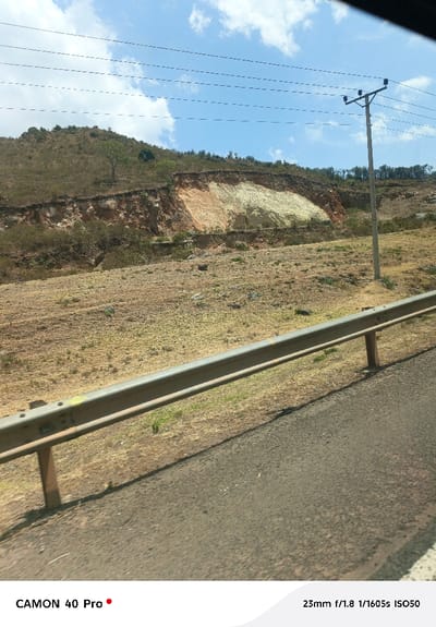 The image displays a daytime outdoor scene located in Gilgil ward, Kenya, characterized by dry, open terrain. In the immediate foreground, an asphalt road surface with a white edge line runs diagonally from the bottom right. A metallic, two-bar guardrail with vertical support posts borders the road on the left, separating it from a dry, light brown, sparsely vegetated embankment.

The midground consists of a broad, arid field covered with dry, yellowish-brown grass and dirt, with scattered small rocks and minimal green foliage. A single light-gray utility pole, equipped with multiple horizontal cross-arms and power lines, stands upright in this field towards the right side of the frame, extending wires horizontally across the upper sky.

The background is dominated by a hillside featuring a significant exposed section of earth, indicative of erosion or excavation. This exposed area exhibits distinct strata of reddish-brown, light orange, and a prominent pale yellow/off-white material. The upper reaches of the hillside are covered with sparse, dry vegetation and scattered green trees. The sky above is clear blue with dispersed white cumulus clouds, traversed by several dark, thin overhead wires. No human individuals, animals, or active subjects are visible within the frame.