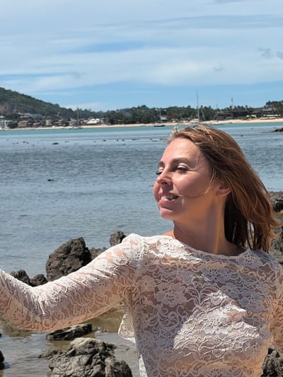 A medium shot shows a Caucasian woman in her late 20s or early 30s from the chest up, standing on a rocky shore with the ocean and a distant coastline in the background. The woman is facing to her right, with her head turned slightly towards the camera, and is smiling. Her eyes are closed, and her hair, which is light brown and windswept, frames her face. She is wearing a white, lace, see-through blouse. The background shows a calm, blue body of water, with a shoreline featuring buildings and lush green hills. Several sailboats are visible on the water. The scene appears to be in a tropical location, possibly Baan Bo Phut, Thailand, given the coastal environment and vegetation. The lighting suggests it is daytime, with bright sunlight casting shadows. The rocks in the foreground are dark and uneven.