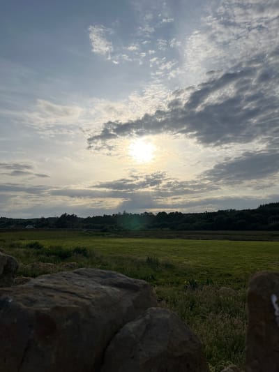 The image captures a scenic view of a grassy field in Terhillion, Ireland. The setting appears to be an outdoor landscape, possibly a rural area, with a stone wall in the foreground partially obscuring the view of a lush green field. Beyond the field, a dense line of trees forms the horizon, and a bright sun shines through a sky filled with scattered clouds. The sun's rays create a dramatic and ethereal effect, casting a soft glow over the scene. The overall impression is one of natural beauty and tranquility, with the time of day appearing to be late afternoon or early evening, given the angle of the sun and the soft light. There is no visible text in the image.
