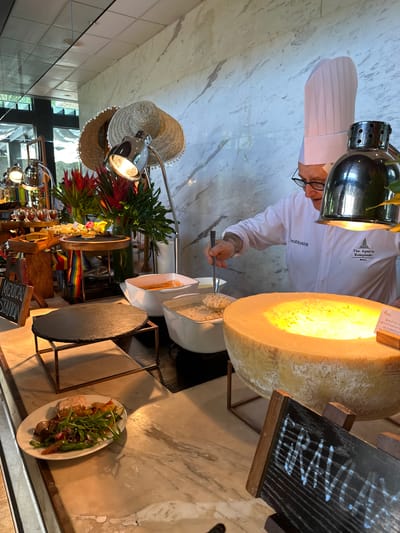 The image depicts a chef in a white uniform and chef's hat, preparing risotto in a large Parmesan cheese wheel at The Apurva Kempinski Bali in Nusa Dua, Indonesia. The cheese wheel is lit by a heat lamp above. The scene is set on a marble counter, with a chalkboard menu displayed nearby. Other food items, including a plate of salad and a "Mexican Station", are visible in the background.