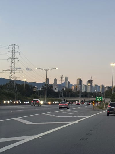 This is a photograph of a highway in Burnaby, Canada, likely during dusk. The sky is a pale blue with hints of orange near the horizon. The highway has multiple lanes of asphalt with white lane markings. Several cars are driving on the highway, with their headlights on. In the background, there is a city skyline with numerous high-rise buildings, some of which appear to be under construction. A large electricity transmission tower is visible on the left side of the image, and trees and hills are also present in the distance. A green highway sign indicating "EXIT 37" with an arrow pointing right is visible on the right side of the image. The overall scene depicts a suburban or urban highway environment.
