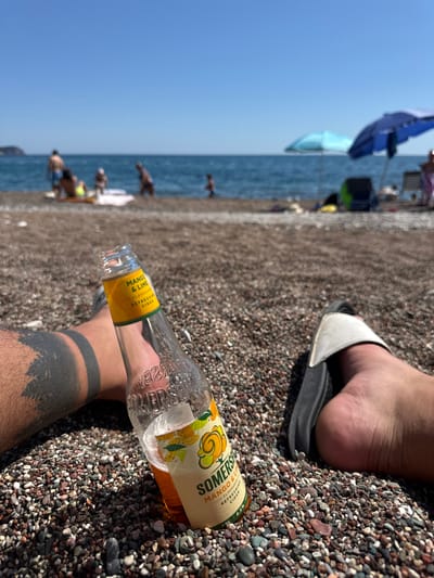 The image captures a first-person perspective on a pebbly beach in Rafailovići, Montenegro, under a clear, bright sky. In the immediate foreground, a person's legs and feet are visible, resting on the multi-colored pebble surface. The left leg displays a prominent black band tattoo, suggestive of a forest or mountain landscape at its base. A partially consumed Somersby Mango & Lime flavored refreshing cider bottle, with its yellow and green label clearly legible, rests upright on the pebbles between the person's legs. The right foot wears a white-strapped, black slide sandal.
The midground, intentionally blurred, reveals several individuals on the beach near the sea. Visible figures include adults standing and seated, and at least one child. Some are near the water's edge, while others appear to be relaxing on towels or mats. Two beach umbrellas are discernible: one light blue and one dark blue. Beyond them, the Mediterranean Sea extends under a clear, light blue sky. A distant landmass or island is faintly visible on the horizon to the left. The overall scene depicts a sunny day at a populated beach.