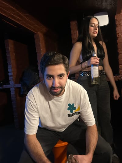 Two individuals are on a brick staircase. A dark-haired woman, lying on her back above, clutches a bottle of wine. A man, wearing a white "Butter" t-shirt, looks directly at the camera, holding a glass. The setting appears to be a dimly lit indoor space, possibly a bar or club, suggested by the brick staircase. The woman is wearing a black top and black jeans with a belt. The man is in dark trousers and an orange seat is partially visible behind him.