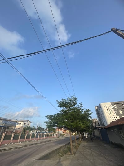 Ilado, Nigeria. A street view shows a road running parallel to a fence. Buildings stand on either side of the road. Overhead, power lines crisscross the clear, blue sky with scattered clouds. A tall, modern building with a white facade and patterned openings stands on the left. A tree with dense green foliage sits along the sidewalk. A building with "OCEAN CREST SCHOOL" sign is visible. People are visible on the sidewalk.