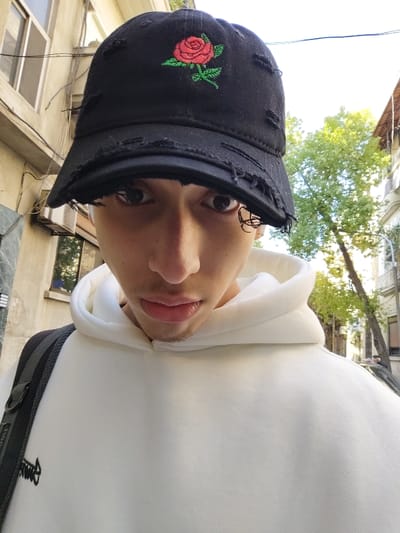 A young person wearing a white hoodie and a distressed black baseball cap with an embroidered red rose. The individual is facing the camera with a slight tilt, showcasing their face. The background reveals an urban setting with buildings and trees, suggesting an outdoor environment. A black strap is visible over the left shoulder.
