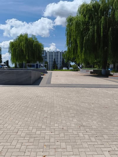 This is a daytime, eye-level, outdoor photograph taken in the city of Ocnița, Moldova. The image features a paved area with herringbone pattern brickwork in the foreground, leading to a darker gray paved area further back. A raised, dark gray stone planter or bench structure is present on the left. Beyond this, there is a walkway with wooden benches visible within white modern structures. Two large weeping willow trees dominate the mid-ground, their green foliage hanging down. In the background, a modern, multi-story white building with vertical window lines stands under a blue sky with scattered white clouds. Several vehicles, including vans and cars, are parked or driving in the background. The overall scene depicts a public outdoor space, possibly a park or plaza, with modern architectural elements and mature trees.