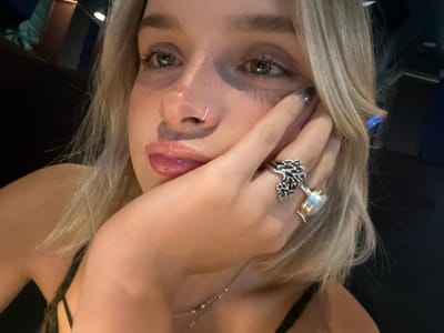 A medium close-up photograph captures a young Caucasian female with blonde hair and light-colored eyes, gazing off-camera to her left. Her right hand supports her chin, with two rings visible: a prominent silver ring featuring an organic, branching design on her ring finger, and a gold-toned ring with a light, iridescent cabochon stone on her middle finger. Her fingernails are coated with a dark, reflective polish. She has a small silver hoop nose ring in her right nostril, and her lips are glossy with a light pink tint. Subtle freckles are present across her nose and cheeks, and a slight shimmer is visible below her eyes. She is wearing a dark, thin-strapped top. The background is dark and extensively blurred, displaying out-of-focus cool-toned ambient light sources, including a distinct blue light in the upper right, consistent with an indoor evening environment such as a restaurant or lounge. The precise location within the city of Victoria, Canada, cannot be determined from the visual information provided in the image.