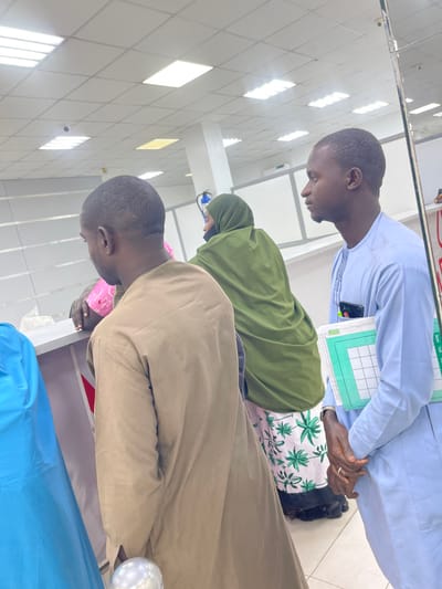 The image depicts an indoor public service facility, possibly in Zaria, Nigeria, characterized by white walls, a white ceiling with square panels, and evenly spaced fluorescent lighting. A black security camera is visible on the ceiling in the upper left.
Three individuals are primarily visible, along with a partial view of a fourth. From left to right:
1. A person in a light blue garment is partially visible, facing right, near a grey-topped counter.
2. A male individual stands with his back to the camera, wearing a light brown, long-sleeved tunic. He leans slightly over the grey counter, with his right arm bent at the elbow and resting on its surface.
3. A female individual stands behind the counter, positioned with her back mostly to the camera, but slightly turned towards the right. She wears a dark olive green hijab covering her head and shoulders, and a white skirt with a repeating green leaf pattern. A pink object is held in her visible right hand.
4. A male individual stands in profile on the right, facing right. He has short dark hair and wears a light blue traditional tunic with embroidered details on the chest. He holds a light green-bordered notebook or folder tucked under his left arm. A dark-colored smartphone is partially visible in a pocket on his right side. His hands are clasped in front of him.
All individuals are standing. The environment suggests a waiting area or a service point. A reflective surface, possibly a mirror or glass partition, is present on the far right edge of the frame. A silvery, spherical object is visible at the bottom center of the image.