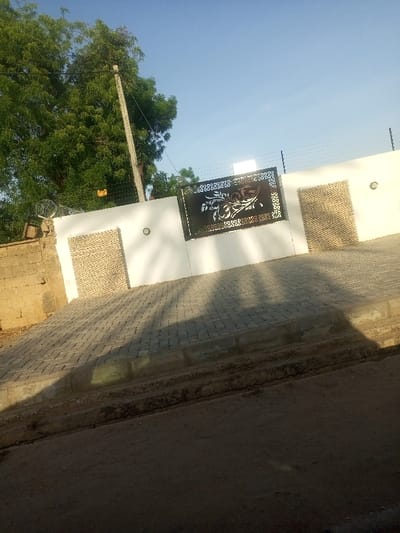 The image shows a white concrete wall with a black metal gate. The gate is decorated with a floral pattern. There are two light fixtures on the wall, one near the gate and another near a door. The door is made of wood with horizontal slats. The wall is built next to a dirt road. There is a concrete sidewalk in front of the wall. The road and sidewalk are covered with shadows. The sidewalk is made of grey bricks arranged in a pattern. There is a tree to the left of the wall, casting a shadow. The scene is taken in Maiduguri, Nigeria, at the coordinates 11.817 latitude and 13.153 longitude.