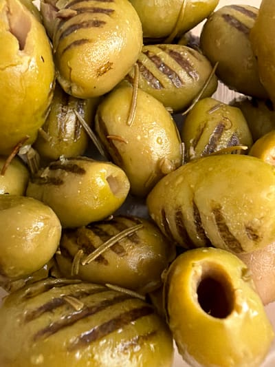 A close-up, overhead shot shows a pile of grilled green olives, some with grill marks. A few rosemary sprigs are interspersed among the olives. The olives are glistening, suggesting they are marinated or in oil. The image is cropped tightly, and the background is not visible. No specific location in Istanbul, Turkey can be determined from the image.