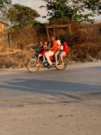 Five people are piled onto a single red motorcycle parked on the edge of an asphalt road. The motorcycle is facing to the left. The person at the front wears a red shirt, and the person at the back wears a red shirt. The middle person wears an orange shirt. Trees and a building can be seen behind the motorcycle. The background also includes some grass, dry bushes, and a small wire fence. The road takes up a large portion of the right-hand side of the image.