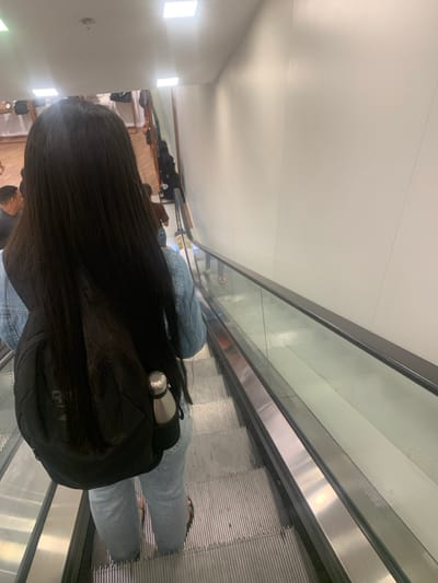 The scene depicts an individual descending an escalator within a commercial establishment in Lauro de Freitas, Brazil. The primary subject, viewed from behind, has long, straight, dark hair and wears a light blue denim jacket over a lighter garment, light-wash jeans, and dark open-toed sandals. A black backpack is carried, with a silver insulated water bottle secured in its right-side mesh pocket. The escalator features corrugated metal steps, transparent glass balustrades, and a black handrail. A circular prohibitory symbol is affixed to the right glass balustrade. To the right, a plain white wall extends upwards, meeting a white ceiling equipped with square recessed light fixtures. In the blurred background on a lower level, other individuals and what appear to be retail displays are discernible.