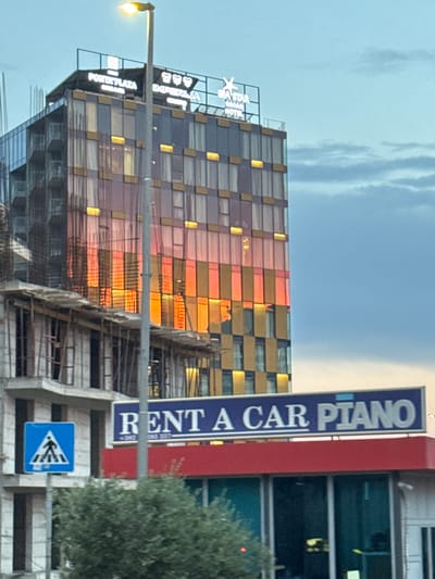 A vertical shot captures a modern building with a glass facade that reflects the warm hues of a sunset. The building's exterior is composed of a grid of rectangular windows, some of which are illuminated with a golden light, creating a striking contrast with the surrounding sky. The sky itself is a mix of soft blues and wispy white clouds, suggesting it is either early morning or late evening.
To the left of the frame, a street lamp with its light on illuminates part of the scene. A tall, vertical blue sign is visible on the right, displaying white text that reads "RENT A CAR PIANO". Adjacent to this sign is a red structure, possibly part of another building or advertisement. Below the "RENT A CAR PIANO" sign, a blue pedestrian crossing sign with a white symbol of people walking is mounted.
A lush green tree is partially visible on the right side, adding a natural element to the urban landscape. The building appears to be under construction in some areas, with scaffolding and exposed structural elements visible. Signs on the building, though partially obscured, seem to indicate names like "PORTOPLAZA" and "IMPERIA". The overall impression is that of a contemporary urban environment, likely a commercial or hotel district, with the vibrant sunset adding a dramatic and picturesque quality to the scene. The location is identified as Rafailovići, Montenegro.