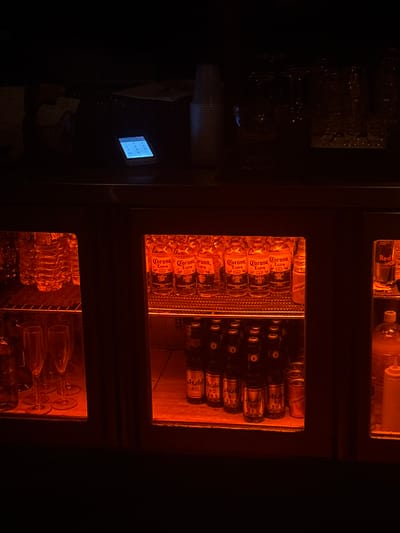 Here is a forensic description of the image:

The image shows a dark interior setting, seemingly a bar or restaurant, with refrigerated shelves displaying beverages. The shelves are illuminated with a warm orange light, highlighting bottles of Corona Extra beer and Asahi beer, among others. There are also bottles of Red Bull and Schweppes tonic. The shelves appear to be a part of a larger refrigerated unit. 

To the left, out of focus, are stacks of white cups and containers, along with what appears to be a digital display with a blue screen. 
Without more visual data, it is not possible to provide precise location details within Hong Kong.