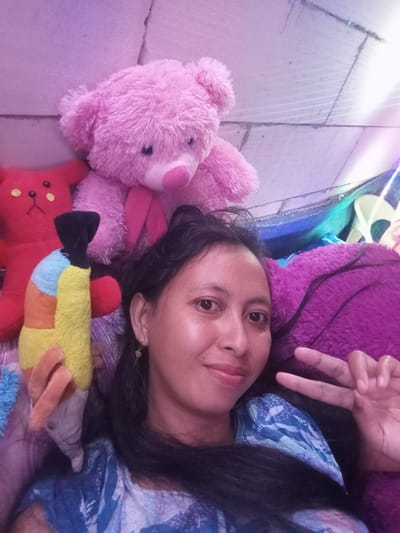 An indoor shot taken in Baron, Indonesia, depicts a woman resting comfortably amidst several plush toys. She is seen from the chest up, lying on her left side with her head on a vibrant purple, fluffy pillow or blanket. The woman has long, dark hair and light-brown skin, and is looking towards the camera with a gentle smile. Her right hand is raised, forming a peace sign. She wears a blue patterned garment on her upper body.
To her right and behind her are three distinct stuffed animals. A red teddy bear with yellow button-like features on its front is partially visible. Adjacent to it lies a colorful plush toy resembling a bird, featuring a yellow body, orange wings, a light blue patch, and a black beak. Further right, a pink, fluffy teddy bear with a pink nose is nestled, its head and upper body visible. The background appears to be a light-colored wall or floor, possibly tiled or paneled, with soft purple and pink hues, suggesting ambient or artificial lighting. The image conveys a relaxed and friendly atmosphere. No text is visible in the media.