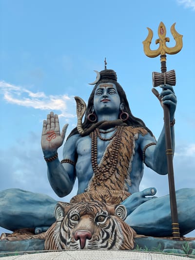The image depicts a large, blue-skinned statue of the Hindu deity Shiva, prominently featuring a trident, a hand adorned with a symbol, and a tiger. The statue is set against a backdrop of a bright blue sky interspersed with scattered white clouds. The location is identified as Pokhara, Nepal.