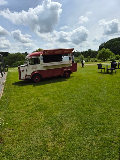 The image shows a white and red Citroen H van, likely converted into a mobile bar or food truck, parked on a grassy field. The van is marked with the name "LET'Z SPRITZ MOBIL", with the word "LET'Z" vertically stacked. A person is seen standing near tables and chairs set up on the grass, further back in the scene. The background includes green trees and a partly cloudy sky. The scene suggests a casual outdoor event. The location is Lenningen, Luxembourg.