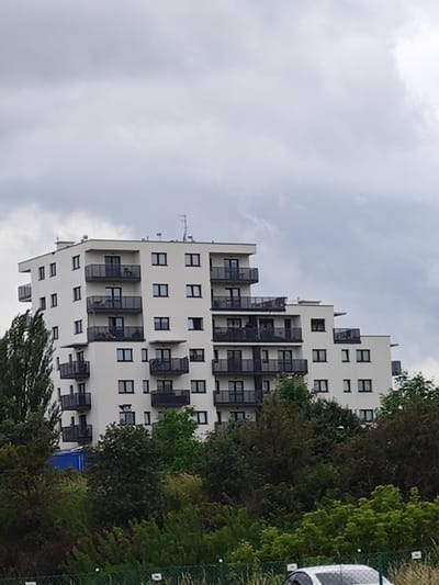 The image shows a modern, multi-story apartment building with a stepped facade, located in Poznan, Poland. The building is white with dark balconies and numerous windows. Trees and foliage surround the base of the building, with a cloudy sky overhead. A small portion of a white car and a fence are visible in the foreground.