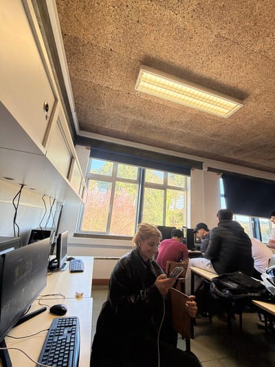 This image depicts an interior scene of a computer lab or classroom located in Vila Real, Portugal. The ceiling is composed of corkboard panels, with a single rectangular fluorescent light fixture providing artificial illumination. Along the left wall, white overhead storage cabinets are mounted. Large windows on the back wall offer a view of exterior trees, some displaying reddish-pink blossoms and others green foliage, under natural daylight; blue blinds are partially drawn along the top of these windows.
The room contains multiple white desks, each equipped with a black desktop computer, monitor, keyboard, and mouse; associated electrical and data cabling is visible. In the foreground, a female with blonde hair, wearing a black jacket, is seated on a wooden chair. She is looking down at a silver smartphone in her hands, which is connected to a white charging cable, and exhibits a smiling facial expression. In the mid-ground and background, several male individuals are present. One male in a red shirt is seated, facing a computer monitor. Another male, wearing a black puffer jacket and a black baseball cap, is similarly positioned in front of a computer. A third male, also in a black puffer jacket, is observed from behind, seated on a chair or desk in the mid-ground right. A white crumpled paper object rests on a desk in the foreground left, near a computer setup. Additionally, a black backpack and a white motorcycle helmet are discernible on a desk or chair in the right foreground.