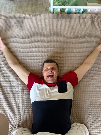An adult male is depicted lying supine on a light brown, horizontally ribbed blanket in a domestic setting within Gornyi (Dvurechenskoe S/P), Russia. He is wearing a short-sleeved tri-color t-shirt (red, white, and dark blue/black with a chest pocket) and light-colored pajamas adorned with small red patterns. His arms are extended outward and upward. His mouth is open wide, exposing his teeth, indicative of a loud vocalization or laughter. In the upper background, light-colored flooring is visible. To the upper right, a section of a white wooden crib is present, containing a green patterned sheet, a light pink pillow, and a blue rectangular object.