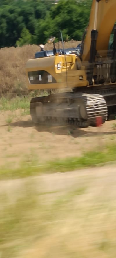 The image shows a close-up of a yellow excavator. The excavator is on a dirt ground with some sparse green vegetation. In the background, there are trees and a hill. The image appears to be taken from a moving vehicle due to the motion blur. There is no discernible indication of the specific location within Waiblingen, Germany, or any people or interactions.