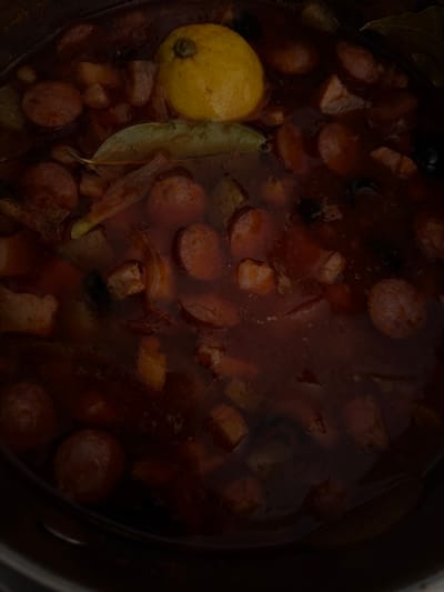 An overhead view depicts the contents of a dark cooking pot, located in Novosaratovka, Russia. The pot contains a dark reddish-brown liquid broth with various solid ingredients submerged and floating. A whole, light yellow lemon with a dark stem is positioned in the upper-central portion of the frame, partially submerged. Below the lemon, a single olive-green bay leaf floats on the liquid surface. Numerous circular slices of reddish-brown sausage are visible throughout the pot, along with irregularly shaped pieces of light brown meat. Scattered dark, oval-shaped objects, consistent with olives, are also present. The scene is dimly lit, with the dark rim of the pot visible along the edges. No individuals are present.