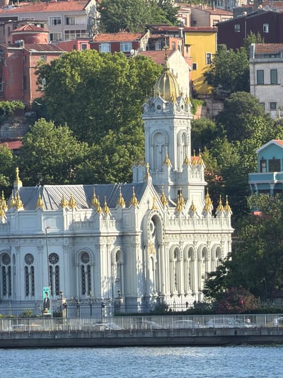 A long shot photograph captures a white church with gold accents situated on a hill overlooking a body of water. The church features a prominent golden dome and numerous smaller gold-capped spires adorning its roofline. Intricate architectural details, including arched windows and ornate carvings, are visible on the church's facade. Lush green trees surround the building, partially obscuring residential structures with red-tiled roofs in the background. A fence separates the church from the water, where several cars are visible on a raised roadway. A banner with the text "KIP" and a man in a tuxedo is displayed on the church's exterior. The water in the foreground reflects the bright sunlight. This is the Bulgarian Orthodox Church of St. Stephen in Istanbul, Turkey, located on the shores of the Golden Horn.