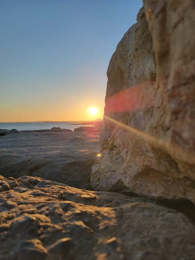 A low-angle shot captures a sunset over the ocean, viewed from a rocky shore. In the foreground, large, textured rocks dominate the frame, their surfaces catching the warm, golden light of the setting sun. A prominent rock face rises on the right, with a bright lens flare streaking across it. In the mid-ground, more rocks extend towards the water. The ocean is calm, with a faint horizon line visible, and a distant landmass with a few structures can be seen. The sky transitions from a clear, pale blue at the top to a warm orange and yellow gradient near the horizon, indicative of sunset. The overall scene is serene and natural, typical of the coastal areas of Quarteira, Portugal.