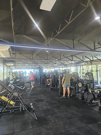 The image shows the interior of a gym, possibly in Miraflores, Peru. The gym is equipped with various weight machines and exercise equipment. Several people are exercising on the machines. The walls and ceiling are dark, with exposed metal beams and lighting fixtures. The floor is covered with black rubber tiles. The gym appears to be well-lit and organized.