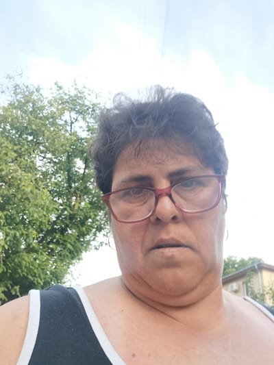 A selfie taken outdoors shows a person with dark, curly hair and glasses with red frames. The person is wearing a black tank top with white trim. Behind the person is a tree with green leaves and a cloudy sky. In the background, a building with windows and an air conditioning unit is visible. The image appears to have been taken in the city of Lomets, Bulgaria.