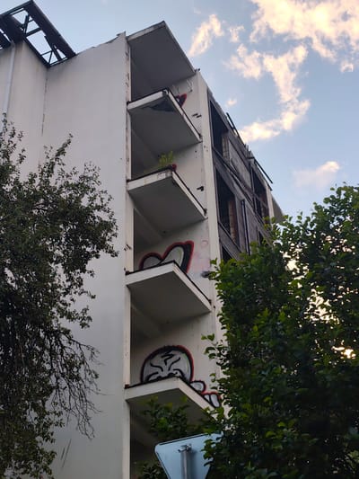 This media file captures a weathered, abandoned multi-story building, likely an apartment complex, in Krasnaya Polyana, Russia. The building exhibits signs of decay, with damaged balconies, exposed structural elements at the top, and wooden boards covering some of the windows. Graffiti art, primarily in red and black, adorns several balconies, depicting stylized faces or abstract shapes. Green foliage from surrounding trees partially obscures the structure, suggesting a natural or overgrown environment. The sky above is partly cloudy, with visible patches of blue and white clouds, hinting at daytime, possibly late afternoon or early evening due to the soft lighting. The overall mood conveyed is one of neglect and urban decay, juxtaposed with the resilience of nature. No people are visible, and no text is discernible in the image.