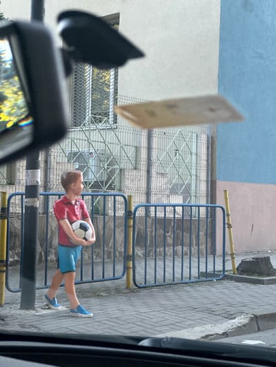 In Dobrich, Bulgaria, a statue of a young boy stands on a paved sidewalk, holding a soccer ball. The boy is wearing a red polo shirt and blue shorts. He is positioned near a blue metal fence and a lamppost. The background shows a building with a light-colored facade and a blue section, along with a metal fence covering a window. A blurry object, possibly a piece of paper, is visible in the upper right portion of the image. The scene is captured from inside a vehicle, with the car's interior visible in the foreground and a reflection of trees in the rearview mirror.