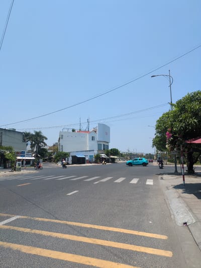 The image depicts a street scene in Tuy Hòa, Vietnam. A light blue pickup truck is driving on the right side of the road, heading away from the camera. There are two motorcycles in the distance. The street has yellow double lines indicating a no-passing zone and white stripes marking a pedestrian crossing. Buildings line the left side of the street, with a modern white building closer to the camera and older, more traditional structures farther away. A street light is visible on the left. Power lines stretch across the sky. The sky is clear and blue. Trees and vegetation are present on both sides of the street.