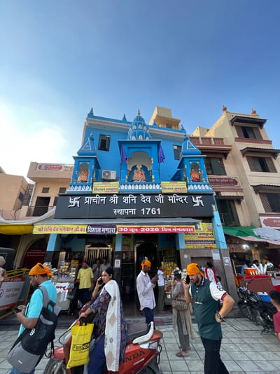 The image depicts the facade of the Shri Shani Dev Mandir in Amritsar, India. The temple is painted a vibrant blue with intricate architectural details, including domes and decorative spires. A prominent black sign above the entrance displays Hindi text and the year 1761. Several individuals are visible in the foreground and middle ground. Some are wearing orange turbans, a common religious garment. People are engaged in various activities, such as talking on mobile phones, looking at their phones, and carrying bags. The street in front of the temple appears to be a busy marketplace, with stalls selling goods and motorcycles parked nearby. The sky is clear blue, suggesting it is daytime.