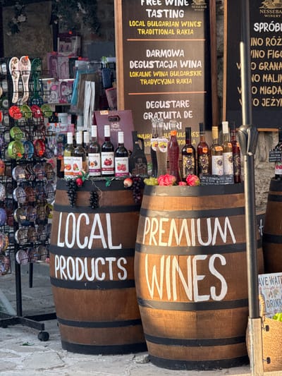 This image depicts a street scene in Nesebar, Bulgaria, focused on a display of wines and local products. Two large, wooden wine barrels are prominent, with white lettering painted on them. The top barrel reads "PREMIUM WINES," and the bottom barrel says "LOCAL PRODUCTS." Both barrels are filled with bottles of wine, some with decorative labels featuring images of fruit like pomegranates and peaches. Small bunches of grapes and artificial roses are placed as decorative elements around the bottles.

Below the barrels, a rack displays various souvenirs, including flip-flops with "BULGARIA" written on them, decorative plates, and small bowls adorned with intricate patterns. Some of these items are priced, with a "30€" tag visible.

To the left of the barrels, a black sign lists "FREE WINE TASTING" and "LOCAL BULGARIAN WINES TRADITIONAL RAKIA" in English, Polish, and Bulgarian. Another sign, partially visible at the top, also seems to advertise wine, with text in a language that appears to be Bulgarian.

The scene suggests a commercial setting, likely a shop or a market stall catering to tourists. The lighting indicates it is daytime, and the overall atmosphere appears to be bustling and inviting, encouraging visitors to sample local offerings. The presence of both wine and traditional Bulgarian souvenirs points to a cultural and commercial hub.