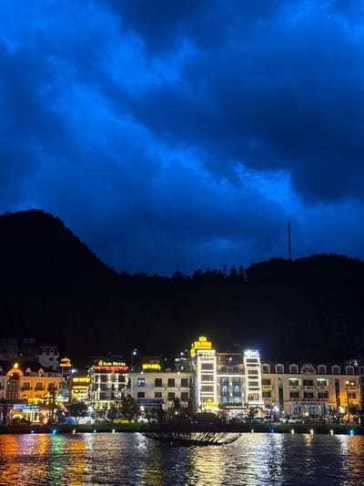 Captured at night, the image presents a body of water reflecting the illuminated cityscape of Sa Pa Ward, Vietnam. The right side of the image is dominated by the light-streaked surface of the water. A line of brightly lit buildings extends from the water's edge, with some appearing to be hotels. Beyond the buildings, the image transitions to a dark, silhouetted mountain range. The left side is filled with a dark blue sky with clouds. A tower, possibly a transmission tower, is visible on the silhouette of the mountain range.